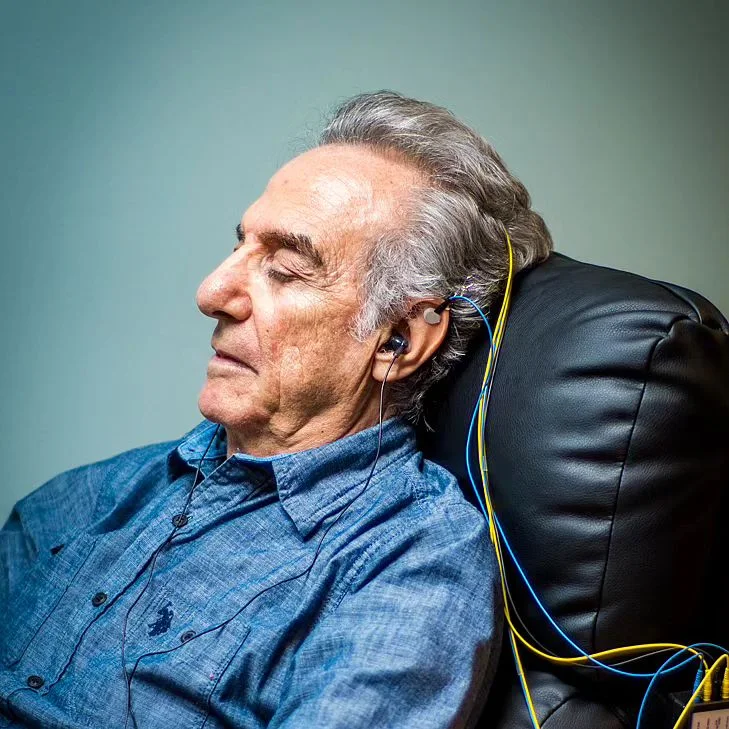 A patient relaxes in a recliner with his eyes closed during a neurofeedback session, sensors clipped to his ears
