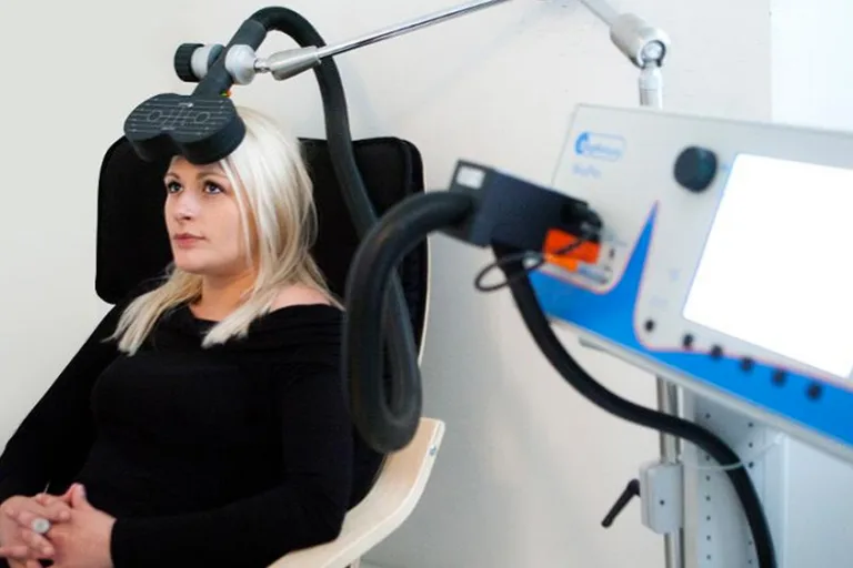 A woman sits relaxed and awake in a TMS chair while the magnetic coil rests on her head